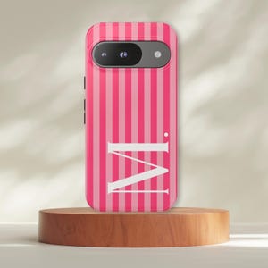 Könnte beinhalten: Rosa und hellrosa gestreifte Handyhülle mit dem weißen Buchstaben "M". Die Handyhülle befindet sich auf einem Holzständer. Die Handyhülle hat eine Kamera und einen Blitz.