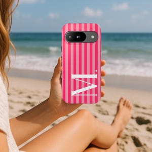 Könnte beinhalten: Rosa und weiß gestreifte Handyhülle mit dem weißen Buchstaben "M". Die Handyhülle wird vor einem Strandhintergrund mit dem Meer im Hintergrund gehalten.