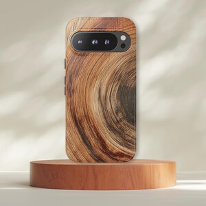 May include: A wooden phone case with a natural wood grain pattern. The case is brown with a dark center and a camera lens. The phone case is displayed on a small wooden pedestal.