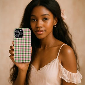 May include: A smartphone case with a green, pink, and cream plaid pattern. The case has a black border and a camera cutout. The phone case is held by a person with long, dark hair and a light pink top.