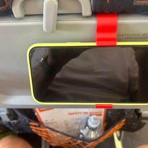May include: A bright red plastic phone mount hooked onto the back of an airplane seat tray table. The mount holds a smartphone with a neon yellow case in a horizontal position, allowing for hands-free viewing during travel on an aircraft.
