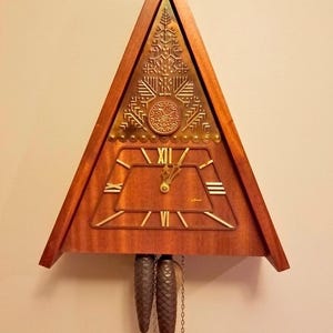 May include: A triangular wooden wall clock featuring a brass plate with embossed tree and bird motifs above a trapezoidal clock face. The face displays gold Roman numerals and hands. Two dark, textured pinecone-shaped weights hang from chains.