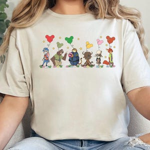 Puede incluir: Camiseta color arena con una ilustraci&oacute;n de dibujos animados de personajes animales sosteniendo globos en forma de coraz&oacute;n. Los personajes est&aacute;n alineados, con estrellas encima. La palabra "SAND" est&aacute; impresa en la parte inferior.