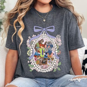 May include: Dark heather gray t-shirt featuring a colorful illustration of cartoon animal characters inside a decorative oval frame with a purple bow and floral accents. The text "DARK HEATHER" is printed at the bottom of the shirt.