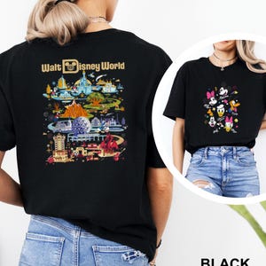 Op de afbeelding: Zwarte T-shirts met Disney World-ontwerpen. E&eacute;n shirt toont een kleurrijke parksc&egrave;ne met de tekst "Walt Disney World". De andere toont Mickey Mouse, Minnie Mouse, Donald Duck, Daisy Duck en Goofy. Het woord "BLACK" is ook zichtbaar.