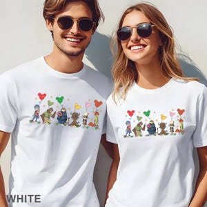 Puede incluir: Camisetas blancas con un dise&ntilde;o de dibujos animados colorido que presenta varios personajes de animales sosteniendo globos en forma de coraz&oacute;n. La palabra "WHITE" est&aacute; impresa en la parte inferior. Las camisetas las usan dos personas.