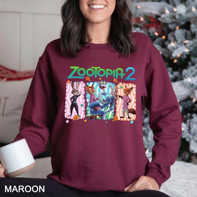 May include: Maroon sweatshirt featuring the text "ZOOTOPIA 2" in green and blue. The graphic includes cartoon characters in rectangular frames with pink and orange accents. The sweatshirt is a solid maroon color.
