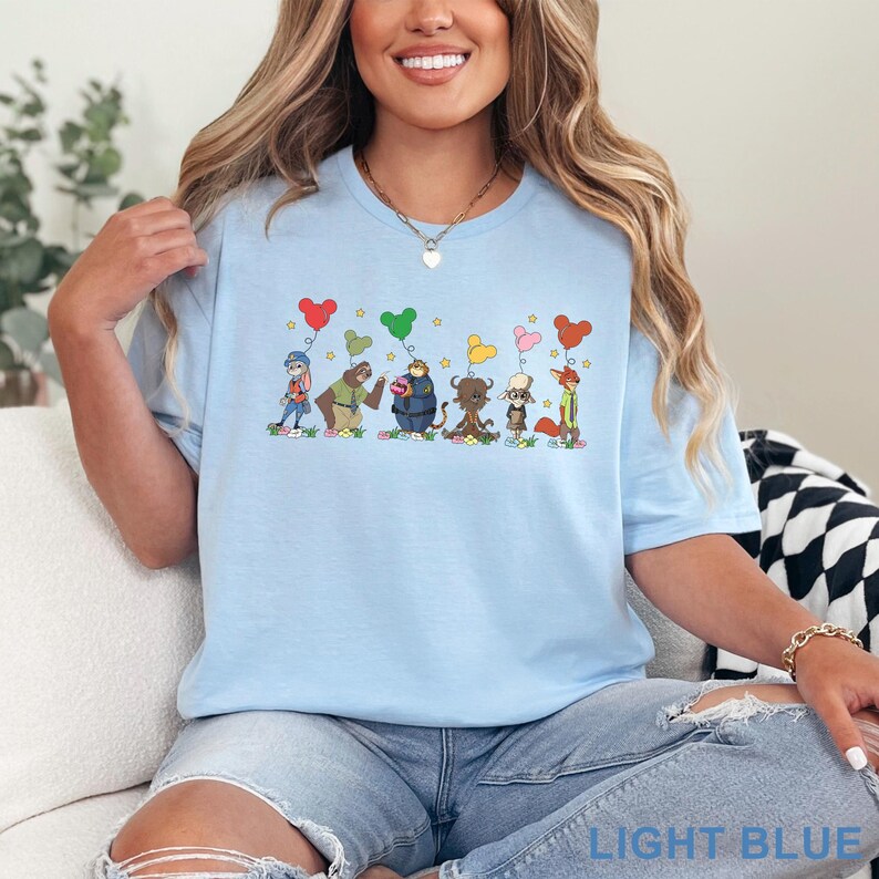 Puede incluir: Camiseta azul claro con una ilustraci&oacute;n caprichosa de animales de dibujos animados sosteniendo globos en forma de coraz&oacute;n. El dise&ntilde;o incluye un conejo, un oso, un mapache, un toro, un perro y un zorro. El texto "LIGHT BLUE" est&aacute; abajo.