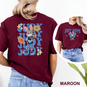 K&ouml;nnte beinhalten: Burgunderfarbenes T-Shirt mit einer Cartoon-Illustration von Judy Hopps aus Zootopia. Das Design zeigt Judy Hopps mit einer Kaffeetasse, einer Karotte und dem Text "Judy". Das Shirt hat auch ein Polizeiabzeichen.
