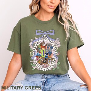 May include: Military green t-shirt featuring a cartoon illustration of various animal characters within a floral and pearl-bordered oval. The design includes a purple bow at the top. The text "MILITARY GREEN" is at the bottom of the shirt.
