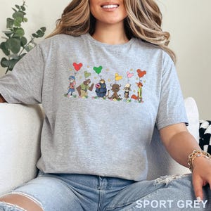 Puede incluir: Camiseta gris deportiva con un dise&ntilde;o de dibujos animados de personajes de animales sosteniendo globos en forma de coraz&oacute;n. Los personajes est&aacute;n de pie sobre hierba con flores. El texto "SPORT GREY" est&aacute; impreso en la parte inferior.