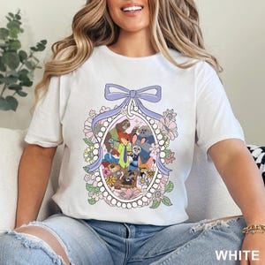 May include: White t-shirt featuring a colorful cartoon illustration of various animal characters within an oval frame, surrounded by flowers and a purple bow. The word "WHITE" is visible at the bottom right.