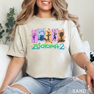 May include: Sand-colored t-shirt featuring a colorful graphic of characters from Zootopia 2. The design includes Judy Hopps, a snake, Nick Wilde, a rabbit, and a water buffalo. The text "ZOOTOPIA 2" is in green and blue.