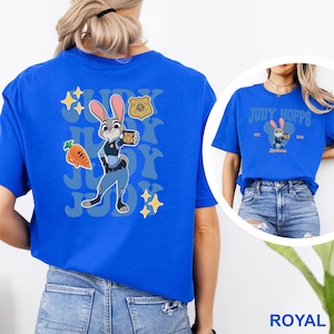 K&ouml;nnte beinhalten: K&ouml;nigsblaues T-Shirt mit Judy Hopps aus Zootopia. Auf der R&uuml;ckseite des Shirts ist ein Cartoon-Bild von Judy Hopps, einer Karotte und einem Polizeiausweis zu sehen. Auf der Vorderseite des Shirts steht "JUDY HOPPS" und das Zootopia-Logo.