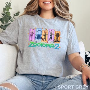 May include: Sport grey t-shirt featuring a colorful graphic from Zootopia 2. The design includes several characters in rectangular frames with the movie title in green and blue. The shirt is made of a soft material.
