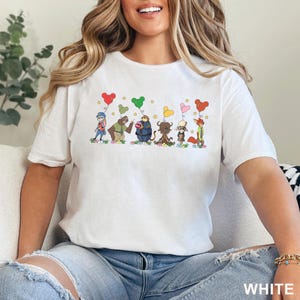 Puede incluir: Camiseta blanca con un dise&ntilde;o de dibujos animados de personajes de animales sosteniendo globos en forma de coraz&oacute;n. El dise&ntilde;o incluye estrellas y la palabra "WHITE" en la parte inferior. La camiseta est&aacute; hecha de un material suave.