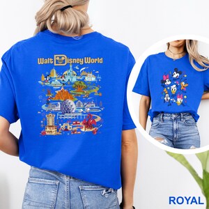 Op de afbeelding: Koningsblauw T-shirt met een kleurrijke afbeelding van Disney World attracties en personages. De achterkant van het shirt toont de woorden "Walt Disney World" in gouden letters boven een gedetailleerde illustratie van het park. Een tweede shirt met Disney-personages is ook zichtbaar.