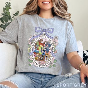 May include: Sport grey t-shirt featuring a colorful cartoon illustration of various animal characters within a floral oval frame, topped with a purple bow. The text "SPORT GREY" is printed at the bottom of the shirt.