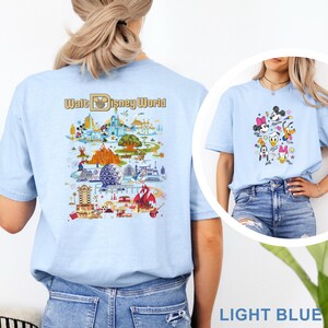 Op de afbeelding: Lichtblauw T-shirt met een kleurrijke afbeelding van Disney-personages en een kaart van Walt Disney World. Het shirt heeft korte mouwen en een ronde hals. De tekst "Walt Disney World" staat op de achterkant.