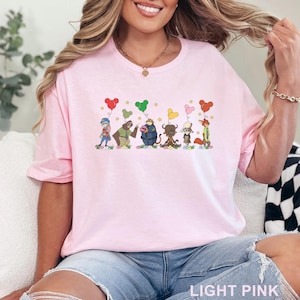 Puede incluir: Camiseta rosa claro con una ilustraci&oacute;n de dibujos animados colorida de varios personajes de animales sosteniendo globos en forma de coraz&oacute;n. La camiseta tiene cuello redondo y mangas cortas. El texto "LIGHT PINK" est&aacute; en la parte inferior.