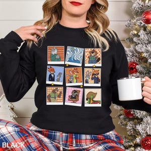 May include: Black crewneck sweatshirt featuring a collage of cartoon character images in polaroid-style frames. The word "BLACK" is printed in white at the bottom. The sweatshirt is worn by a person.