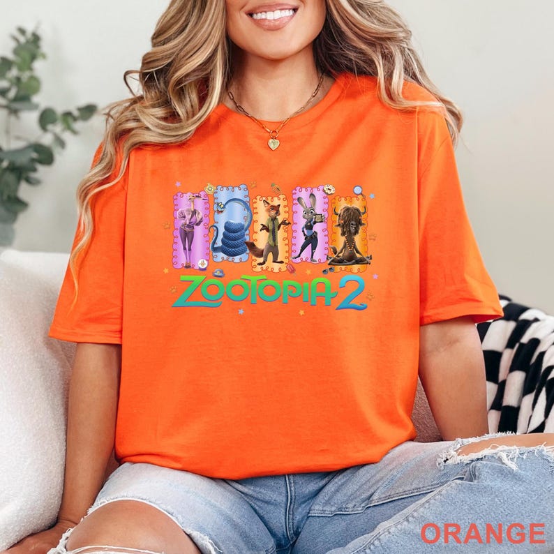 May include: Orange t-shirt featuring the text "Zootopia 2" in green and a colorful graphic of characters from the movie. The shirt is a vibrant orange color.