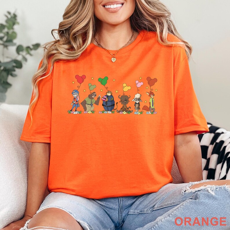 Puede incluir: Camiseta naranja brillante con una ilustraci&oacute;n de dibujos animados de personajes de animales con globos en forma de coraz&oacute;n. La palabra "ORANGE" est&aacute; impresa en la parte inferior. La camiseta es de un material suave.