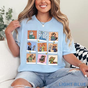 Pode incluir: Camiseta azul claro com uma grade de retratos de personagens de desenhos animados em estilo polaroid. Os personagens s&atilde;o animais antropom&oacute;rficos. A camisa tem as palavras "LIGHT BLUE" impressas na parte inferior.