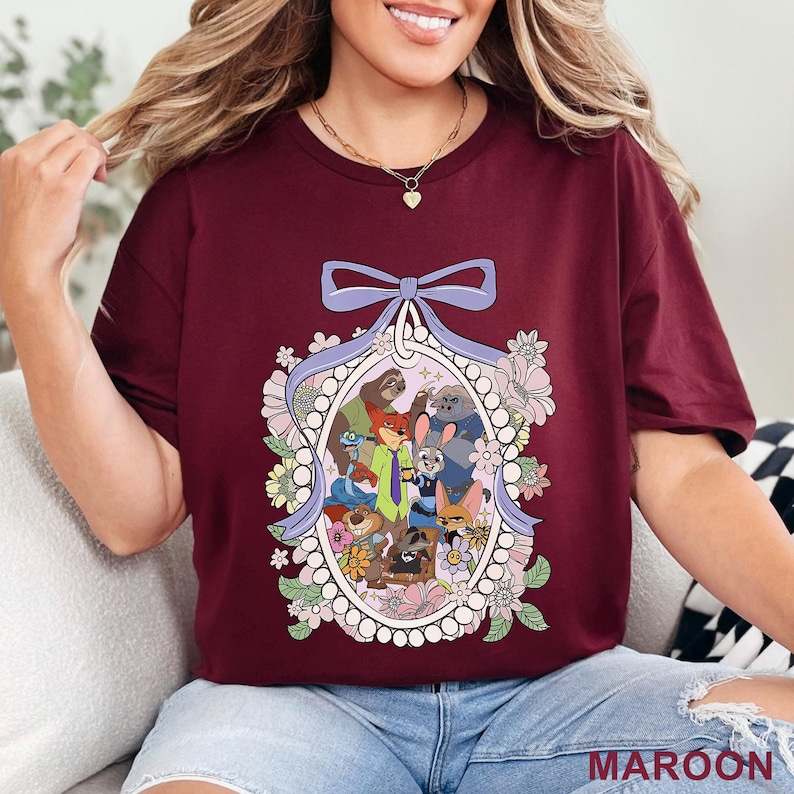 May include: Maroon t-shirt featuring a colorful illustration of animated animal characters within an oval frame, surrounded by flowers and a bow. The word "MAROON" is printed at the bottom right. The shirt is a crew neck style.