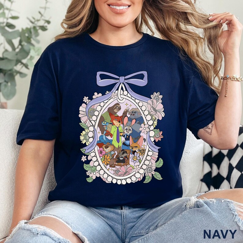 May include: Navy blue t-shirt featuring a colorful cartoon illustration of various animal characters within an oval frame, surrounded by flowers and a purple bow. The word "NAVY" is printed at the bottom right.
