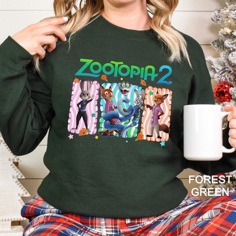 May include: Forest green sweatshirt featuring the text "ZOOTOPIA 2" above a graphic of characters from the movie. The graphic includes Judy Hopps, Nick Wilde, and other characters in colorful frames. A white mug is held in the hand.
