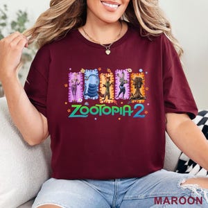 May include: Maroon t-shirt featuring a colorful graphic from Zootopia 2. The design includes several characters in rectangular frames with decorative borders. The text "Zootopia 2" is in a stylized font with paw prints and stars.