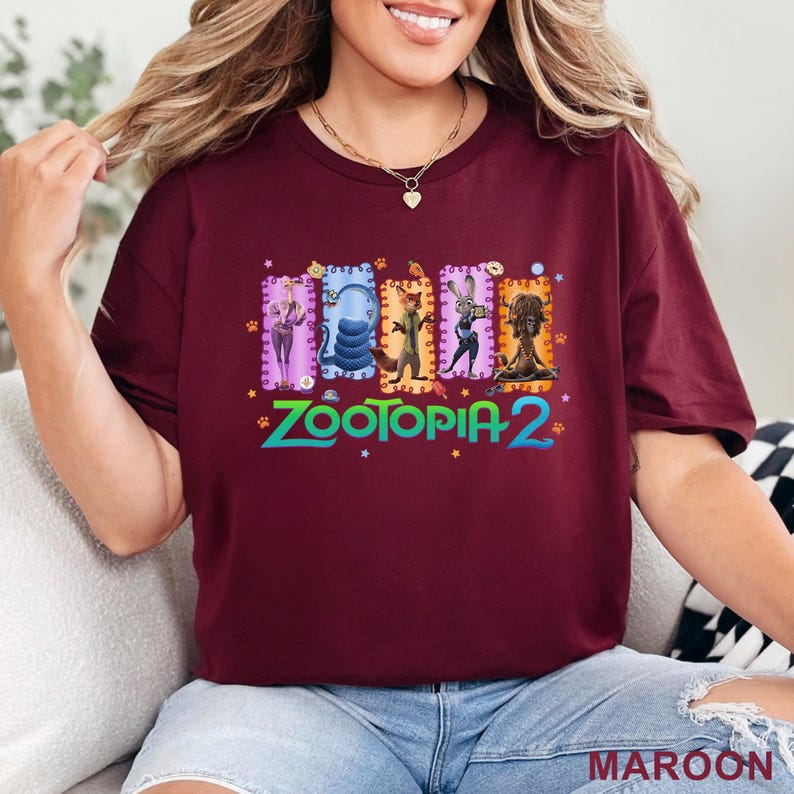 May include: Maroon t-shirt featuring a colorful graphic from Zootopia 2. The design includes several characters in rectangular frames with decorative borders. The text "Zootopia 2" is in a stylized font with paw prints and stars.