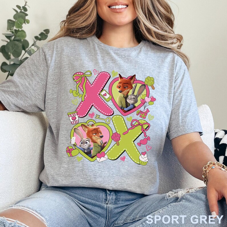 Puede incluir: Camiseta gris deportiva con un dise&ntilde;o de Zootopia. El gr&aacute;fico presenta a Nick Wilde y Judy Hopps, junto con las letras "XO" en rosa y verde. El dise&ntilde;o incluye formas de coraz&oacute;n y elementos decorativos.