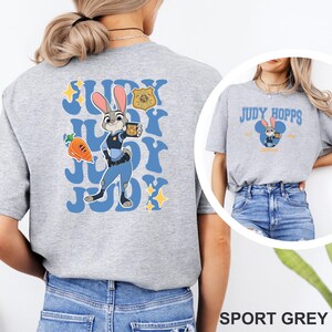 K&ouml;nnte beinhalten: Sportgraues T-Shirt mit einem Cartoon-Bild von Judy Hopps aus Zootopia. Das Design enth&auml;lt den Text "Judy Hopps" und ein Cartoon-Bild von Judy Hopps in ihrer Polizeiuniform. Das Shirt hat auch den Text "Judy" in blauer Schrift.