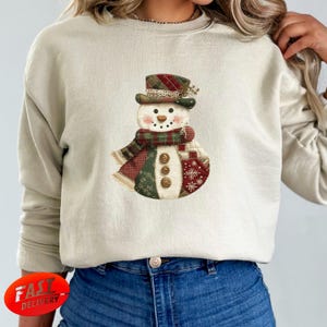 Könnte beinhalten: Beiges Sweatshirt mit einem festlichen Schneemann-Design. Der Schneemann hat einen karierten Hut und Schal, Knöpfe und einen Patchwork-Körper mit Schneeflocken-Akzenten. Ein gemütliches Winter-Themen-Kleidungsstück.