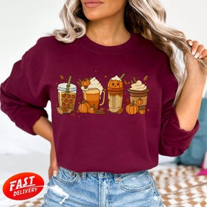 Könnte beinhalten: Burgunderfarbenes Sweatshirt mit einem herbstlichen Motiv von vier Pumpkin-Spice-Getränken. Die Getränke befinden sich in transparenten Bechern und Tassen, mit Schlagsahne, Kürbissen und Blättern. Der Text "FAST DELIVERY" befindet sich in einem roten Kreis.