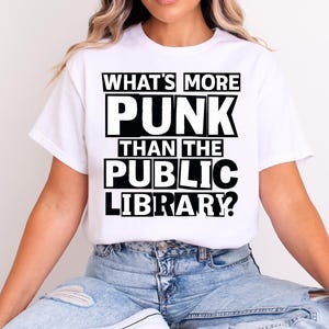 以下が含まれることがあります： 「WHAT'S MORE PUNK THAN THE PUBLIC LIBRARY？」というテキストが黒い太字でプリントされた白いTシャツ。クルーネックのスタイルで、人が着ています。