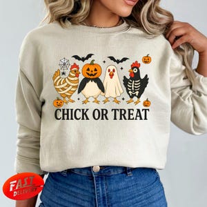 Könnte beinhalten: Beiges Sweatshirt mit einem Halloween-Motiv. Das Design zeigt Cartoon-Hühner, die als Kürbis, Geist und Skelett verkleidet sind, mit dem Text "CHICK OR TREAT" darunter. Fledermäuse, Spinnen und Kürbisse vervollständigen das Design.