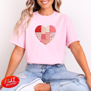 Könnte beinhalten: Ein hellrosa T-Shirt mit einem Patchwork-Herz-Design. Das Herz besteht aus verschiedenen roten und rosa Mustern, darunter Punkte, Streifen und Blumendrucke. Das T-Shirt ist ein lässiger Stil, geeignet für den Alltag.