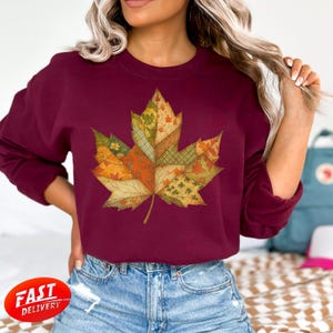 Könnte beinhalten: Ein weinrotes Sweatshirt mit einem großen, farbenfrohen Ahornblatt-Design. Das Blatt besteht aus verschiedenen Patchwork-Mustern in Orange, Grün und Gelb, die ein Herbstthema hervorrufen. Das Sweatshirt hat einen Rundhalsausschnitt und lange Ärmel.