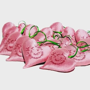 May include: A cluster of pink heart-shaped sachets tied with green ribbons. Each sachet features a printed design in a darker pink. The text on the sachets is in French.