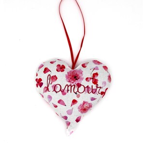 May include: A heart-shaped fabric decoration with a red hanging ribbon. The white fabric is patterned with pink and red flowers and petals. The word "l'amour" is embroidered in red script across the front.