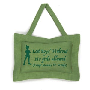 May include: Green fabric tote bag with a rectangular design. The bag features a green silhouette of Peter Pan and the text "Lost Boys' Hideout, No girls allowed (Except Mommy & Wendy)". The bag has a handle for carrying.