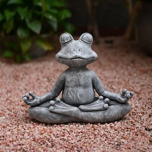 Può includere: Una statuetta di rana con texture in pietra grigia in posizione di meditazione seduta, con gli occhi chiusi e le mani appoggiate sulle ginocchia in un gesto mudra. La rana ha una testa liscia e arrotondata, una bocca larga e piedi palmati.