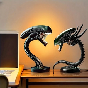 May include: Two Alien-themed lamps with a dark green and black color scheme. The lamps feature the iconic Xenomorph head design, with open mouths revealing internal details. The lamps are on a wooden surface, with a laptop visible in the background.