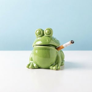 May include: A glossy light green ceramic frog figurine designed as an ashtray. The frog has large, prominent eyes and a wide mouth, with a lit cigarette resting in the corner of its mouth, extending outward to the right side.
