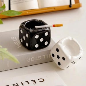 Pode incluir: Dois cinzeiros em forma de dado, um preto com pontos brancos e um branco com pontos pretos, são exibidos. O cinzeiro preto contém um cigarro. Ambos os itens apresentam cantos arredondados e entalhes para apoiar os cigarros.