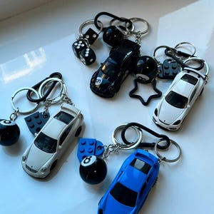 Puede incluir: Llaveros con modelos de coches en miniatura en negro, blanco y azul, junto con dados, una estrella y amuletos de bola de billar. Cada llavero tiene un anillo y un cierre plateado.