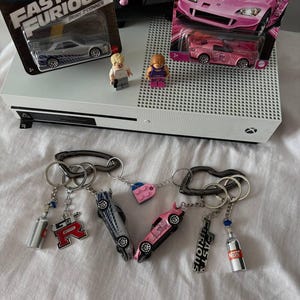 以下が含まれることがあります： シルバーとピンクのFast & Furiousミニカーが付いた2つの金属製キーホルダー。それぞれカラビナに取り付けられています。GT-RとFast & Furiousのロゴチャーム、小さなNOSボトルチャーム、ピンクとブルーのハート型ブロックが付属しています。